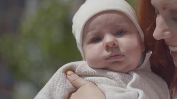 Portrétní zrzavá žena držící dítě v náručí a usmívá se na dvoře zblízka. Dáma si užívá slunného dne s dítětem venku. Mladá matka s dítětem. Šťastná rodina. — Stock video