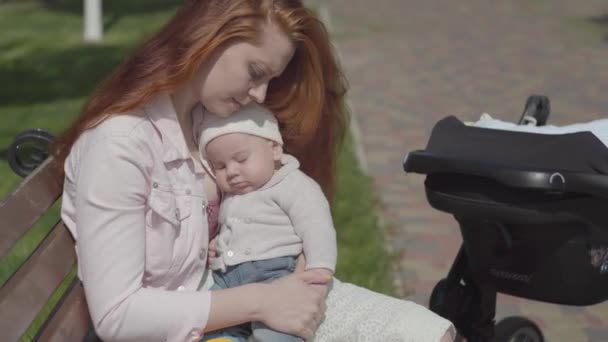 Porträt süße rothaarige Frau, die mit ihrem Kind spielt, das auf der Bank sitzt. der Kinderwagen, der in der Nähe steht. die Dame genießt den sonnigen Tag mit ihrem Baby im Freien. junge Mutter mit Kind. glückliche Familie. — Stockvideo