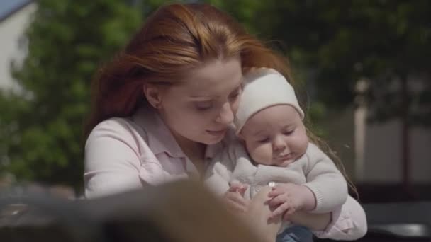 Retrato bastante joven hermosa madre sosteniendo bebé adorable en sus brazos y hablando con él en un día soleado de primavera al aire libre. Familia feliz . — Vídeo de stock