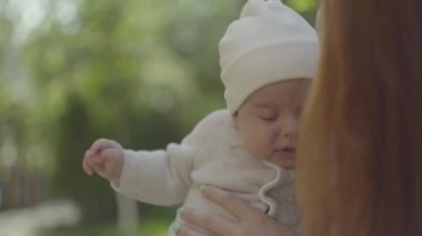 Redhead anne tutarak ve kollarında onu sevimli bebek öpüşme ve bir bahar güneşli bir günde onunla konuşurken açık havada. Mutlu aile.