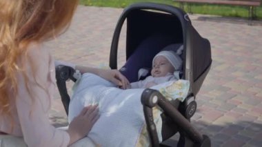 Güzel kızıl saçlı kadın parkta bebek arabasında yatan çocuğuyla oynuyor. Bayan, dışarıda bebeğiyle güneşli bir günün tadını çıkarıyor. Çocuklu genç bir anne. Mutlu aile
