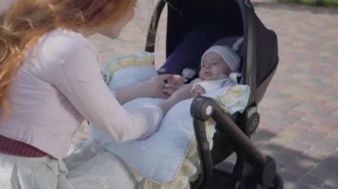 Genç mutlu kızıl saçlı anne arabada bebek ile konuşan ve gülümseyerek ve güzel bir bahar gününde ona bakmak