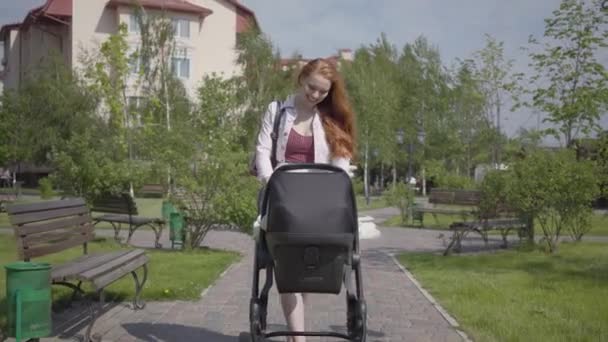 Joven madre pelirroja feliz caminando con carro de bebé y sonriendo a lo largo de la calle en un bonito día de primavera — Vídeos de Stock