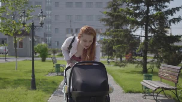 Молодая счастливая рыжая мать ходит с коляской и улыбается вдоль улицы в хороший весенний день — стоковое видео