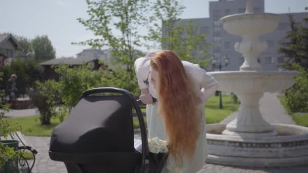 Rozkošná zrzavá matka se naklonila nad kočárek a usmívala se v parku. Roztomilá žena, užívající si slunný den se svým dítětem venku. Mladá matka chodí s dítětem — Stock video