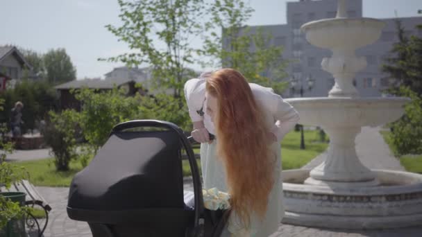 Adorable mamá pelirroja inclinada sobre un cochecito y sonriendo en el parque. Mujer linda disfrutando del día soleado con su bebé al aire libre. Madre joven caminando con un niño — Vídeos de Stock