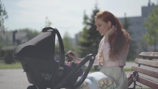 Zijaanzicht van mooie roodharige vrouw spelen met haar kind liggend in de kinderwagen in het Park close-up. De Vrouwe genieten van de zonnige dag met haar baby buitenshuis. Jonge moeder met een kind. Gelukkige familie — Stockvideo