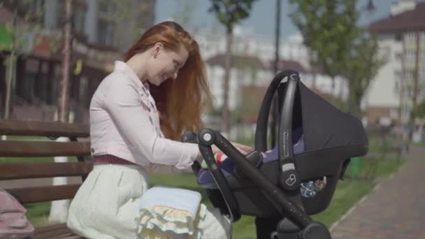 Kızıl saçlı kadın, parkta bebek arabasında yatan çocuğuyla oynuyor. Anne, bebeğiyle güneşli bir günün tadını çıkarıyor. Çocuklu genç bir anne. Mutlu aile — Stok video