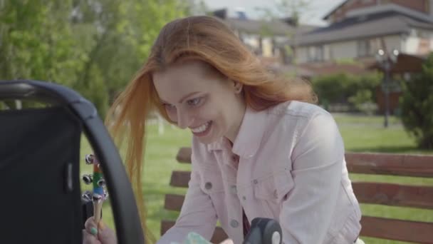 Retrato de bela mulher ruiva brincando com seu filho deitado no carrinho no parque de close-up. A senhora desfrutando o dia ensolarado com seu bebê ao ar livre. Jovem mãe com um filho. Família feliz — Vídeo de Stock