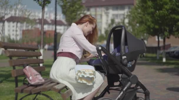 Retrato mulher ruiva brincando com seu filho deitado no carrinho no parque close-up. Mãe desfrutando o dia ensolarado com seu bebê ao ar livre. Jovem mãe com um filho. Família feliz — Vídeo de Stock