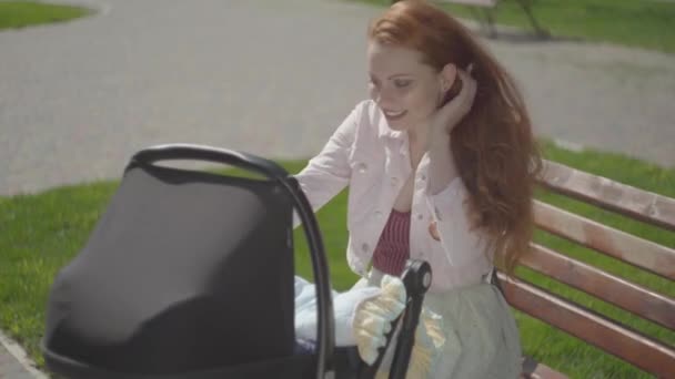 Bankta oturan çocuğuyla oynayan kızıl saçlı bir kadın portresi. Bebek arabası yanında duruyor. Bayan, dışarıda bebeğiyle güneşli bir günün tadını çıkarıyor. Çocuklu genç bir anne. Mutlu aile — Stok video