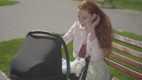 Portret roodharige vrouw spelen met haar kind zittend op de Bank. De kinderwagen staat in de buurt. De Vrouwe genieten van de zonnige dag met haar baby buitenshuis. Jonge moeder met een kind. Gelukkige familie — Stockvideo