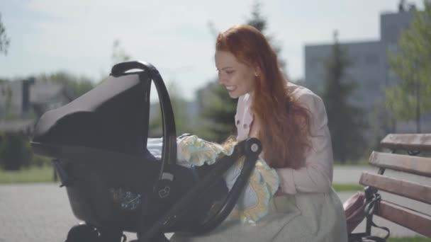 Mladá šťastná zrzka matka sedí na lavičce v blízkosti domu s kočárek a usmívá se a ukolébavka ho na pěkný jarní den — Stock video