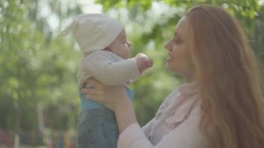 Genç güzel kızıl saçlı anne tutan ve kollarında onu sevimli bebek öpüşme ve bir bahar güneşli bir günde onunla konuşurken açık havada. Mutlu aile.