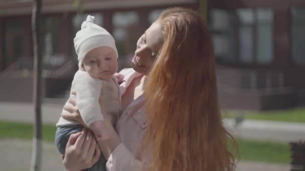 Portre genç sevimli güzel kızıl anne kollarında sevimli bir bebek tutarak ve bir bahar güneşli bir günde onunla konuşurken. Dışarıda çocuğu olan bir anne. Mutlu aile. — Stok video