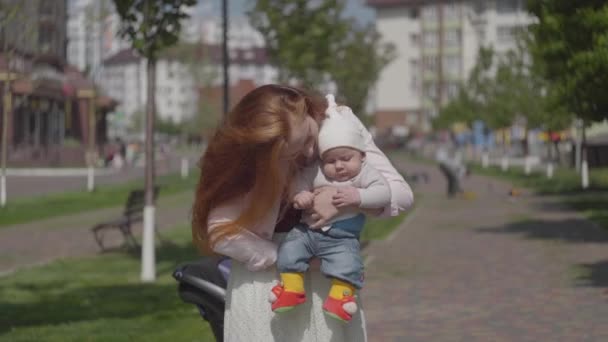 A fiatal, gyönyörű vörös hajú anya egy aranyos kisfiút tart a karjaiban és beszél hozzá egy tavaszi napsütéses napon. Anya egy gyerekkel a szabadban. Boldog család.. — Stock videók