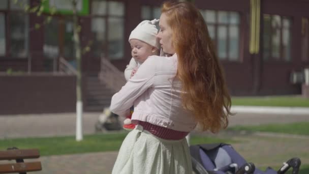 Porträtt av bedårande kvinna som håller barnet i armar och leende på gården närbild. Damen njuter av den soliga dagen med sin unge utomhus. Ung mamma med barn. Lycklig familj. — Stockvideo