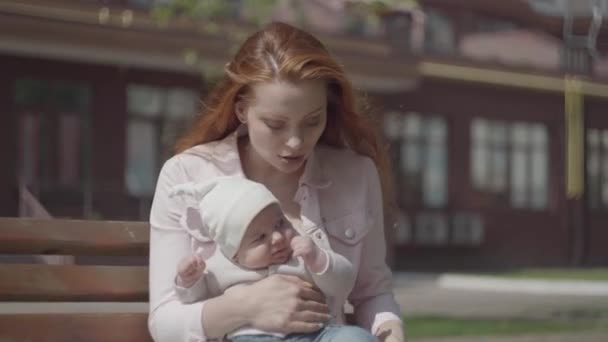 Bankta yakın çekimde oturan çocuğuyla oynayan kızıl saçlı güzel bir kadının portresi. Bayan, dışarıda bebeğiyle güneşli bir günün tadını çıkarıyor. Çocuklu genç bir anne. Mutlu aile. — Stok video