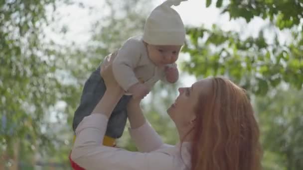 Portret jonge mooie roodharige moeder spelen met haar schattige baby zoon op straat in haar armen in een lente zonnige dag. Gelukkige familie — Stockvideo