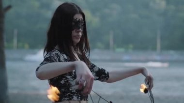 Ağaçların önünde nehir kıyısında duran alev ile bir gösteri gerçekleştiren maskeli sevimli zarafet kadın portresi. Usta fireshow sanatçı akşam ateş hayranları ile dans gösteren. Yavaş çekim