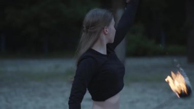Grace blond cute confident woman performing a show with flame standing in the forest or park. Skillful fireshow artist showing mastery of motion of fire in the evening