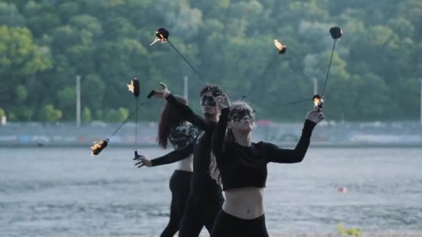 黒い服とマスクを着た巧みな男と2人の女性が、川岸に立ちながら炎と共にショーを行う。夜のジャグリングと火の動きの習得を示す巧みなファイアショーアーティスト — ストック動画