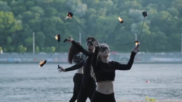 Homme habile et deux femmes en vêtements noirs et masques exécutent un spectacle avec flamme tout en se tenant sur la rive de la rivière. Artistes de feu habiles montrant la maîtrise de la jonglerie et le mouvement du feu dans la soirée — Video