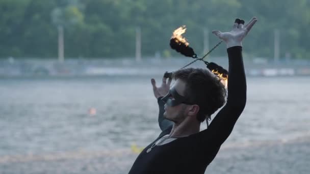 Porträtt skicklig ung smal man i svarta kläder och mask utför en show med flamma stående på Riverbank. Professionell fireshow artist visar behärskning av jonglering och rörelse av eld i — Stockvideo