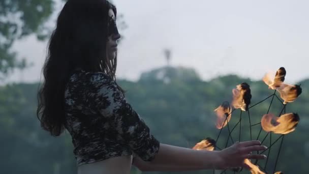 Retrato de jovem mulher graça em máscara realizando um show com chama em pé na margem do rio em frente às árvores. Artista de fogos de artifício habilidoso mostrando dança com os fãs de fogo à noite. Movimento lento — Vídeo de Stock