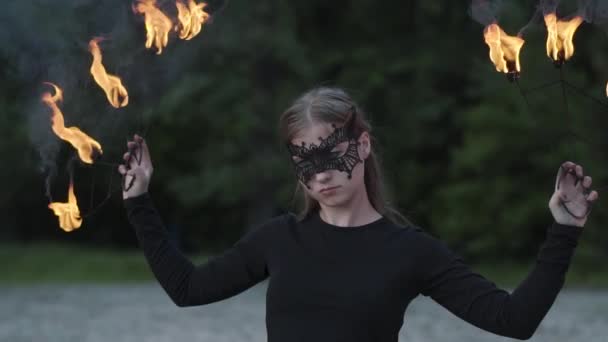 Porträtt av ung vacker kvinna i mask utför en show med flamma framför träd. Skicklig fireshow konstnär som visar dans med eld fläktar på kvällen. Slow motion — Stockvideo