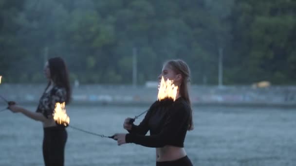 Dos mujeres jóvenes realizando un espectáculo con bolas de fuego de pie en la orilla del río. Hábiles artistas de fuegos artificiales que muestran dominio del movimiento de fuego. Movimiento lento — Vídeo de stock