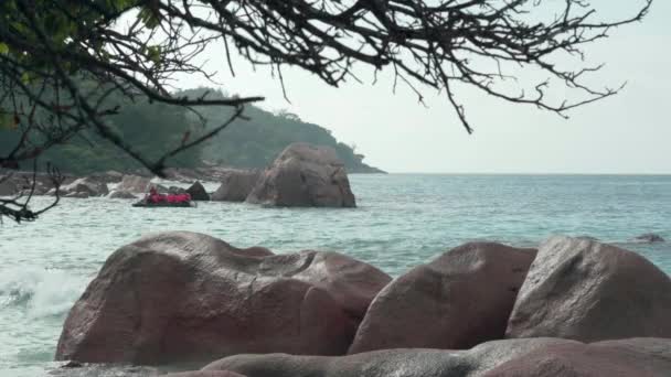 Seychelles. La isla Praslin. La lancha a motor con gente moviéndose en el agua azul del mar en el fondo. Disparando desde detrás de árboles y piedras grandes. Turismo, relax, vacaciones, concepto de viaje — Vídeo de stock