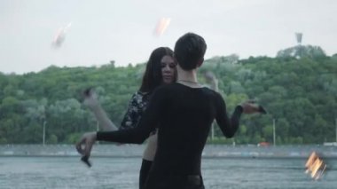 Siyah giysiler genç güzel kız ve yakışıklı adam nehir kıyısında alev ayakta gösterisi performans. Adam başını üzerinde güçlü ateşli Jet exhaling iken ateş topu Golding kadın. Yavaş hareket