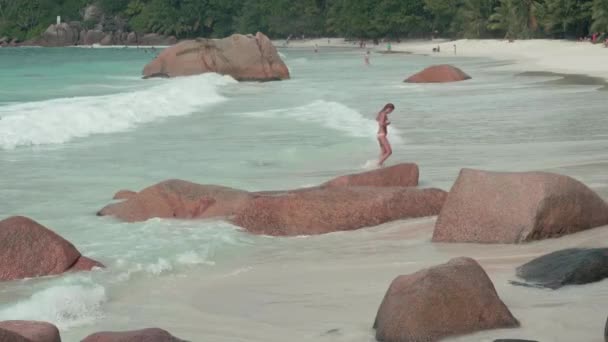 Seicheles. Ilha Praslin. Ondas limpas rolam na praia de areia e grandes pedras deitadas na areia no fundo. Turistas caminhando na praia longe. Turismo, relaxar, férias, conceito de viagem — Vídeo de Stock