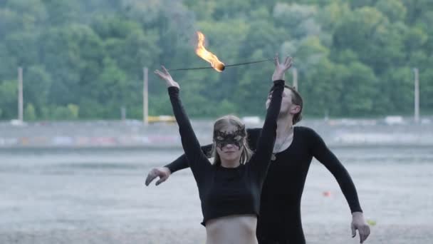 Young beautiful girl and handsome man in black clothes performing show with flame standing on the riverbank. The woman golding fireball while man exhaling powerful fiery jet over her head. Slow motion — Stock Video