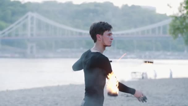 Porträtt av ung man som utför en show med flamma på Riverbank med en bro på bakgrunden. Skicklig fireshow artist som visar behärskning av jonglering och rörelse av eld. Slow motion — Stockvideo