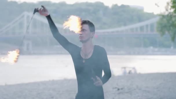 Hombre profesional realizando un espectáculo con llama en la orilla del río con un puente en el fondo. Artista experto en fuegos artificiales que muestra maestría en malabares y movimiento de fuego. Movimiento lento — Vídeo de stock