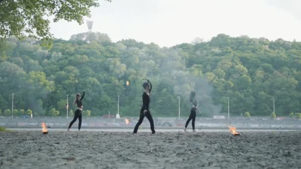 Jovem homem magro e duas mulheres bonitas realizam simultaneamente show com chama enquanto estão em frente à estrada e bosques. Artistas de fogos de artifício habilidosos mostrando domínio do movimento de fogo à noite — Vídeo de Stock