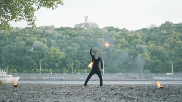 Joven hombre delgado en ropa negra y máscara realizando un espectáculo con llama de pie en la orilla del río. Artista de espectáculo de fuego hábil que muestra el dominio del movimiento del fuego al aire libre — Vídeo de stock