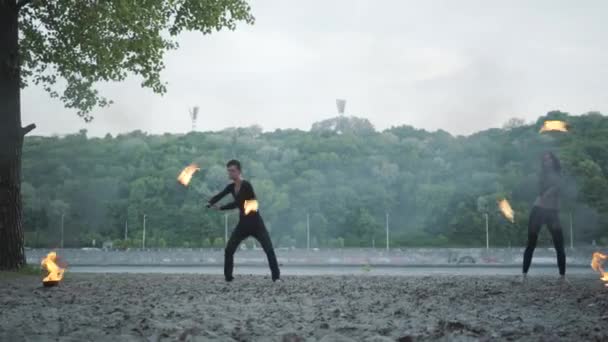 Ung vacker flicka och stilig man i svarta kläder utför show med flamma stående på flod stranden. Kvinnan Golding eld klot medan man andas ut kraftfull eldig Jet över hennes huvud. Slow motion — Stockvideo