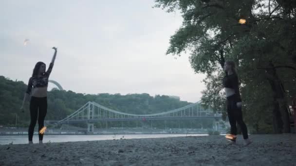 Twee jonge vrouwen die een show met vlam ballen staande op de rivieroever. Bekwame fireshow vrouwelijke kunstenden die beheersing van motie van brand tonen. Onderste weergave — Stockvideo