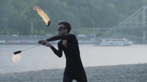 Молодий стрункий чоловік в чорному одязі і масках виконує шоу з полум'ям, що стоїть на березі річки. Умілий художник, що демонструє майстерність руху вогню на відкритому повітрі — стокове відео