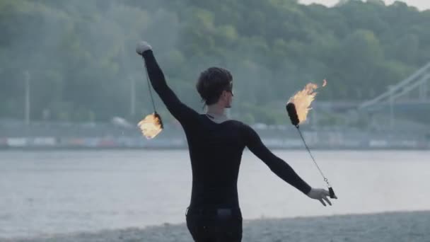 Homem magro jovem em roupas pretas e máscara realizando um show com chama em pé na margem do rio. Artista de fogos de artifício hábil mostrando domínio do movimento do fogo ao ar livre — Vídeo de Stock