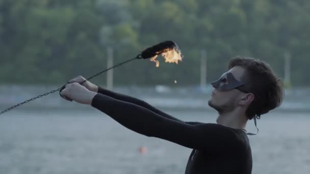 Homem magro jovem em roupas pretas e máscara realizando um show com chama em pé na margem do rio. Artista de fogos de artifício hábil mostrando domínio do movimento do fogo ao ar livre — Vídeo de Stock