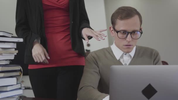 Jonge bescheiden geklede man zittend aan tafel werken op laptop. Hete vrouw met diepe hals bochten over en zet de hand op zijn schouder. De nerd neemt schuchter haar hand af. Secretaresse verleidt baas — Stockvideo