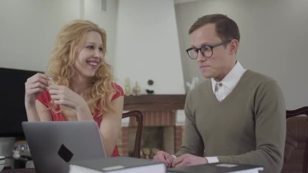 Portrait of cute young man with glasses enthusiastically tells a story of a beautiful seductive girl. Modest nerd and adorable curly blonde indoors. — Stock Video