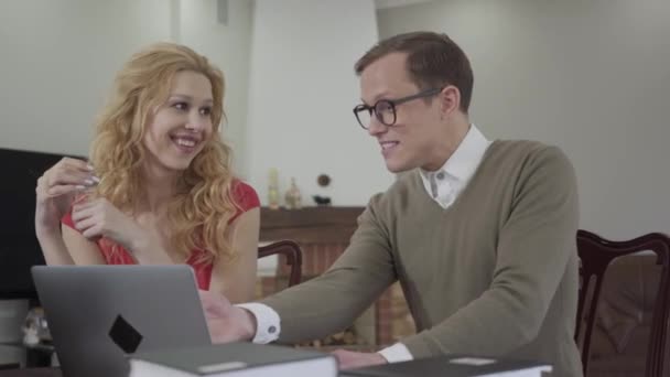 Portrait of cute young man with glasses enthusiastically tells a story of a beautiful seductive girl. Modest nerd and adorable curly blonde indoors. — Stock Video