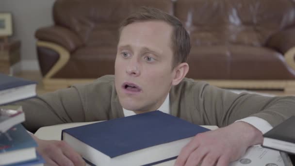 Portrait de jeune homme modestement habillé regardant par derrière le bureau dans le bureau, de nombreux livres sont sur la table. Le nerd frustré et fatigué travaille — Video