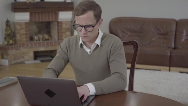 Mladý skromně oblečený muž v brýlích sedícího u dřevěného stolu v kanceláři, který pracuje na svém přenosném počítači. Nerozpoznatelná rozzlobená žena přivezená a Házená do psacího zásobníku knih — Stock video