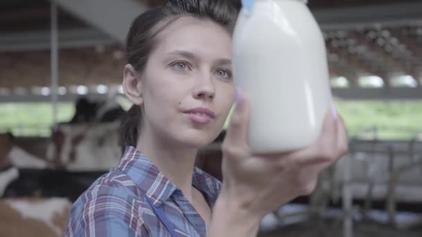 Retrato de una joven trabajadora en la granja de vacas comprobando la calidad de la leche en el primer plano de la botella. El agricultor positivo controla el proceso de trabajo. Industria agrícola, agricultura y ganadería — Vídeo de stock